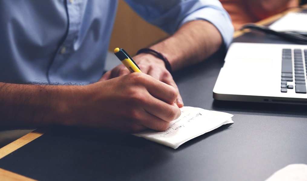 Una persona con camisa azul escribe con bolígrafo amarillo y negro en un pequeño bloc de notas en un escritorio junto a un ordenador portátil abierto, posiblemente documentando detalles relacionados con el acoso laboral en el lugar de trabajo.