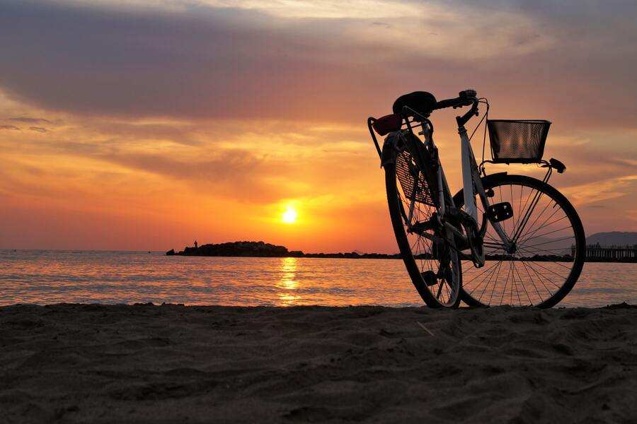 Una bicicleta está aparcada en una playa de arena con el caballete bajado, silueteada contra una puesta de sol naranja y amarilla sobre aguas tranquilas, mientras los pensamientos de 'puedo negarme a firmar el divorcio' perduran en la apacible escena junto a un pequeño saliente rocoso.