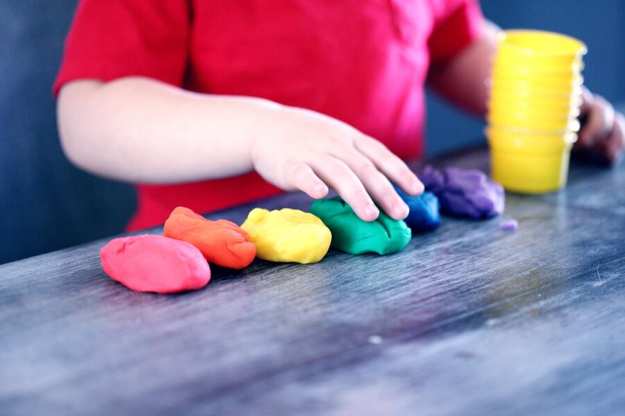 Un niño con camisa roja toca una hilera de coloridas piezas de plastilina -roja, naranja, amarilla, verde, azul y morada- alineadas en una mesa cerca de una pila de vasos de plástico amarillos, mientras los adultos debaten sobre el delito impago de alimentos.