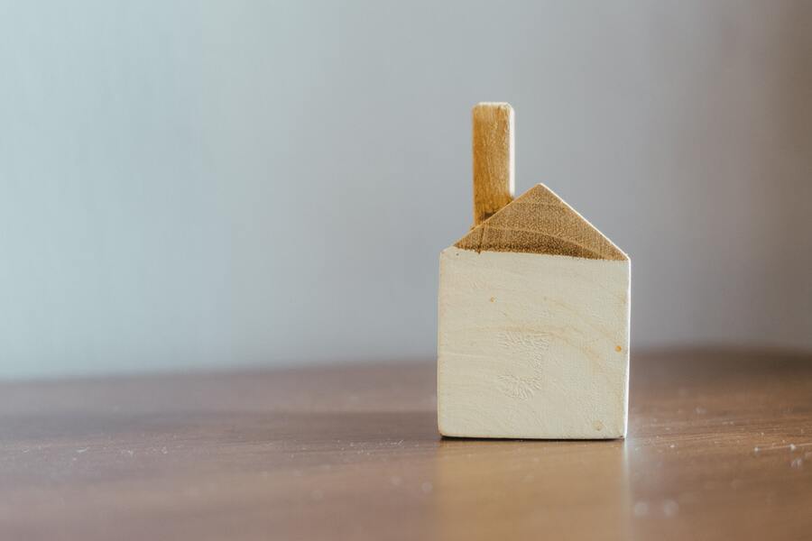 Un pequeño bloque de madera con forma de casa sencilla con chimenea, que simboliza la herencia yacente, se asienta sobre una superficie de madera sobre un fondo liso de color claro.